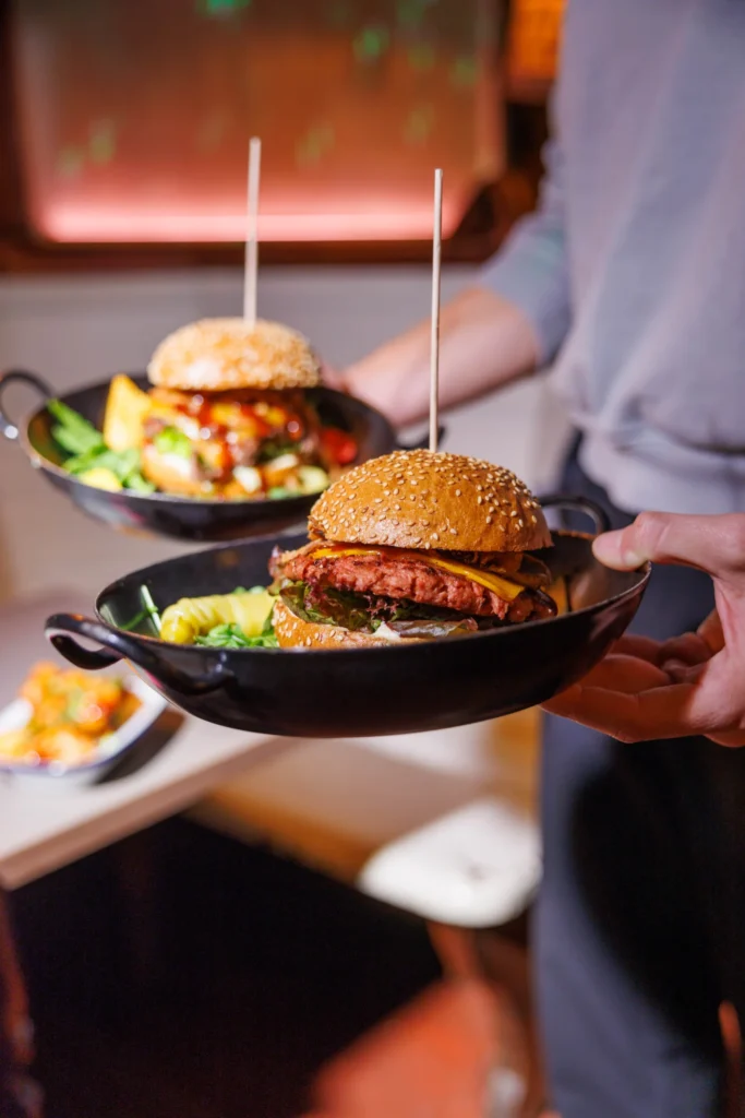 Server carrying burgers and salad in black pans at Let's Be Frank Vienna